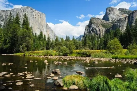 Rocky mountain valley with river, pine trees, and granite cliff formations under blue sky.