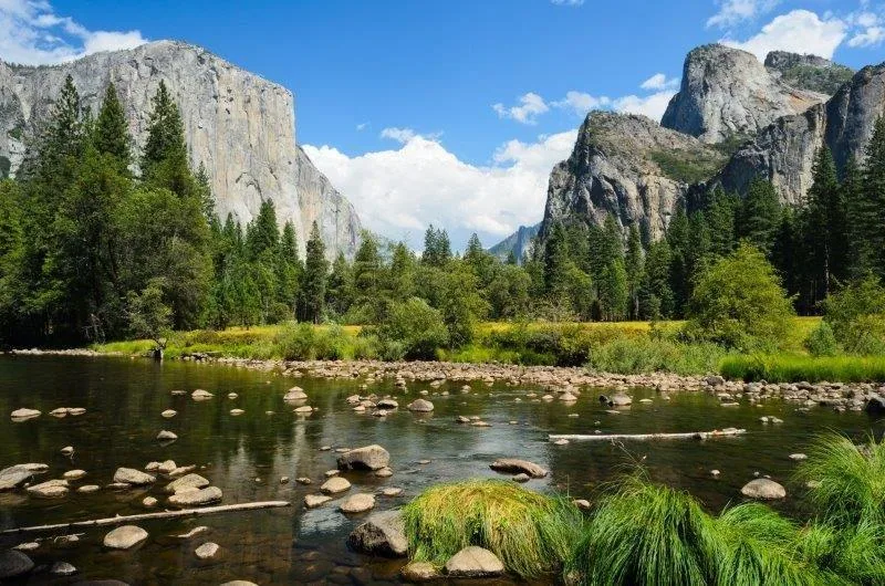 Rocky mountain valley with river, pine trees, and granite cliff formations under blue sky.
