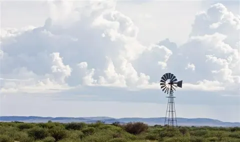 Windmill standing in arid bushland with puffy white clouds and mountains on horizon